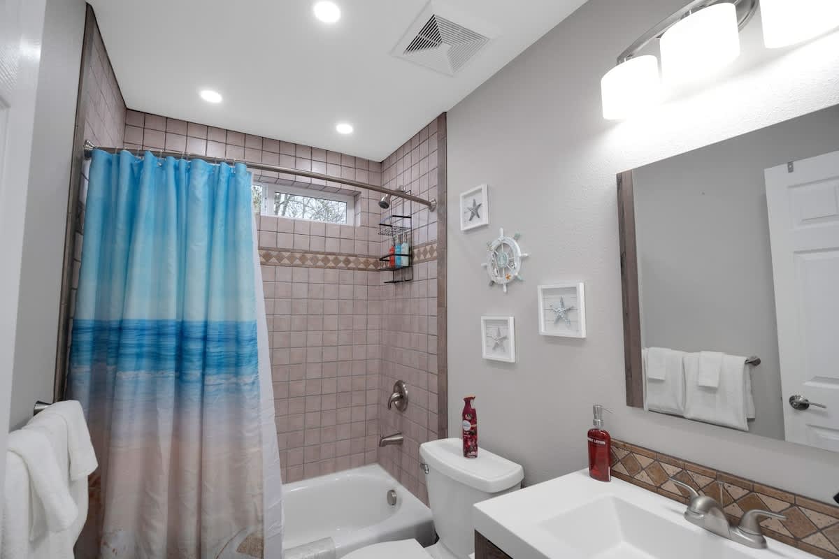 A compact bathroom featuring a shower-tub combo with a blue and white ocean-themed curtain, beige tiled walls, a white toilet, and a vanity with a mirror, all adorned with nautical-themed wall decor.