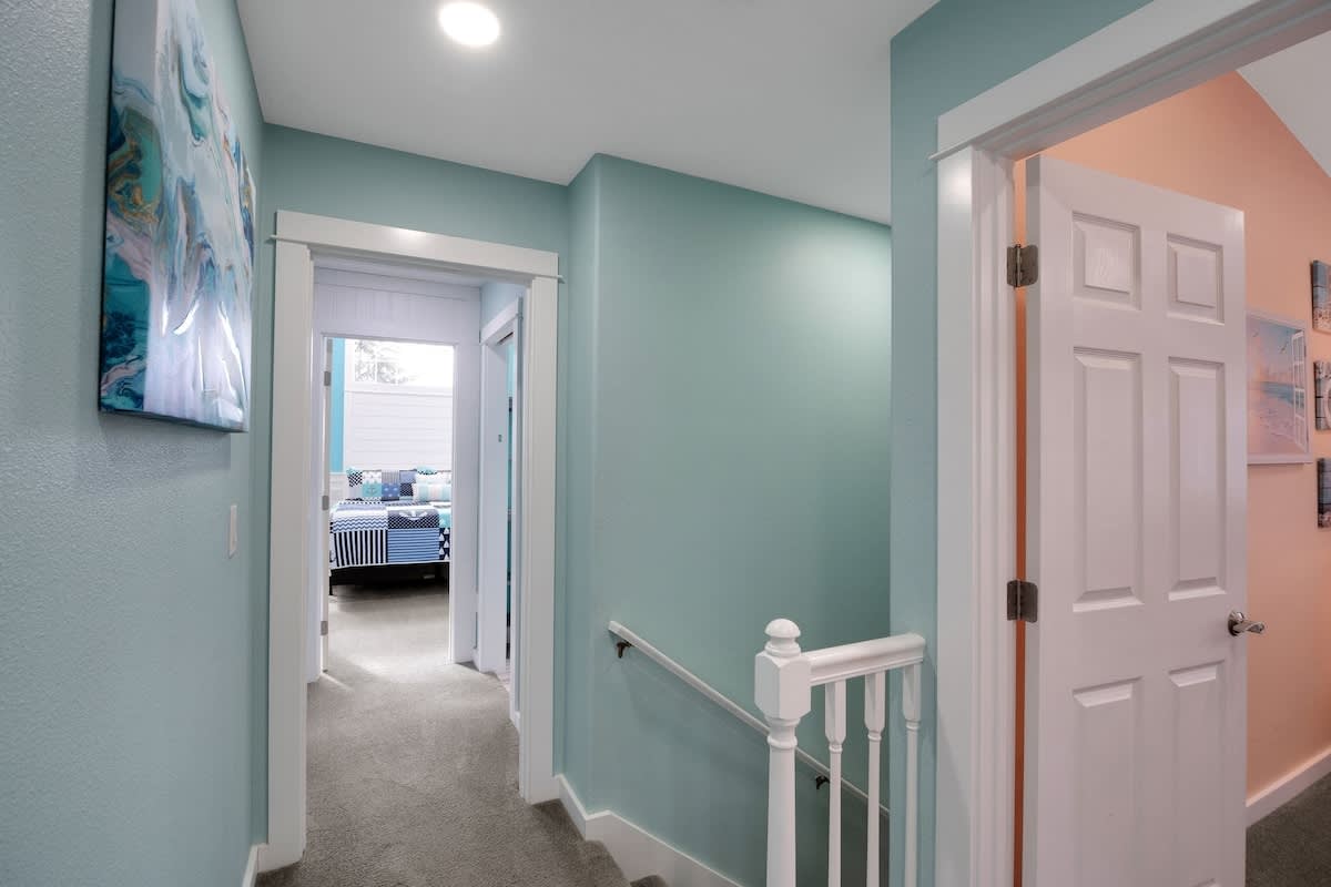 A light blue hallway with white trim and a carpeted floor, leading to several rooms. A white banister is visible on the right, indicating a staircase.