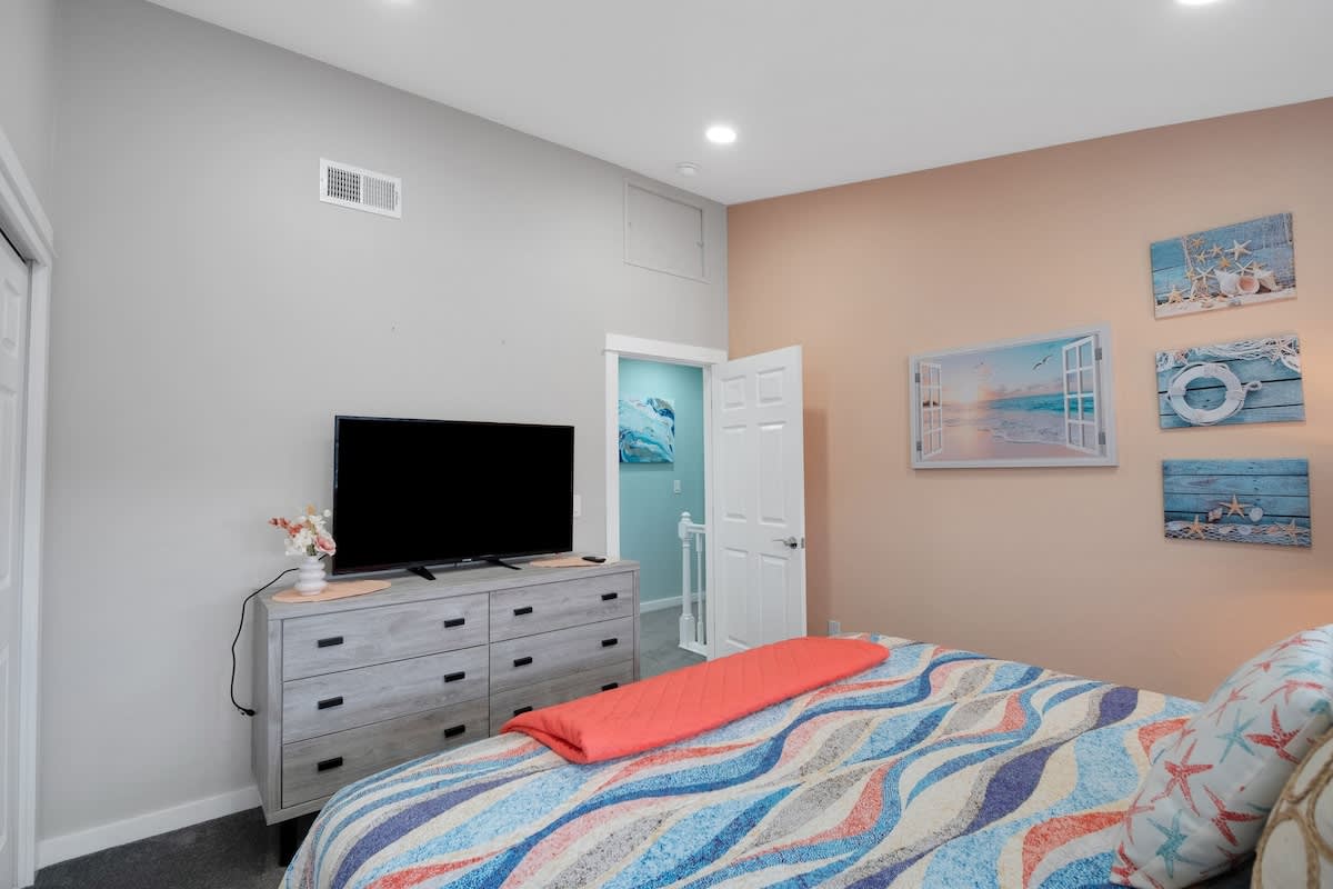 A different view of the peach-walled bedroom, showing a gray dresser with a flat-screen TV, the colorful bed, and a doorway leading to another area. Nautical wall art continues the theme.