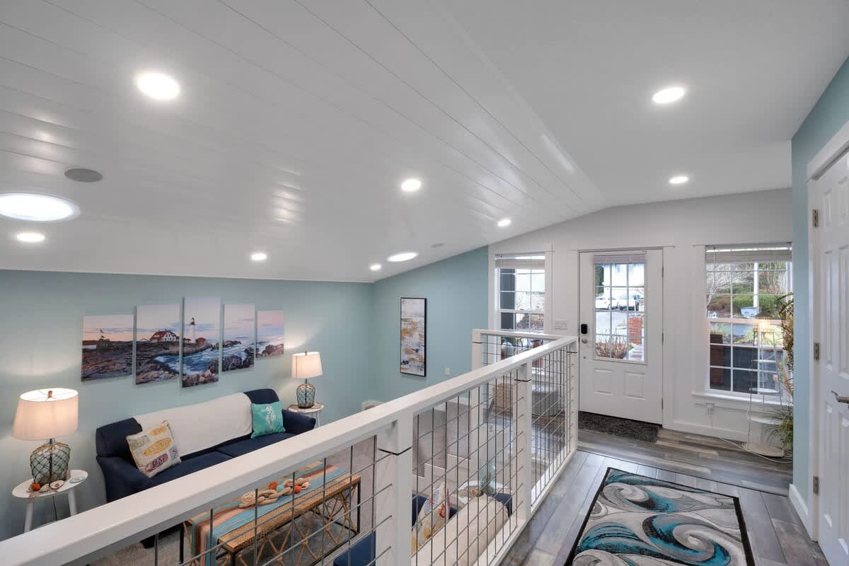 Elevated view of a lower living area with light blue walls and a white shiplap ceiling, featuring a blue sofa, coastal artwork, and an exterior door with windows, seen from a landing with a modern railing.