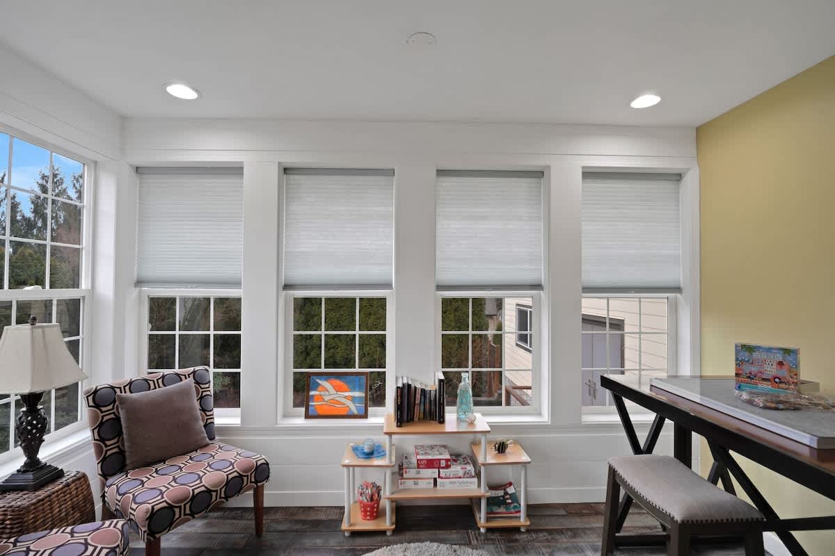 Bright room with multiple large windows providing ample natural light, featuring a patterned armchair, a small bookshelf with games, and dark wood-look flooring.