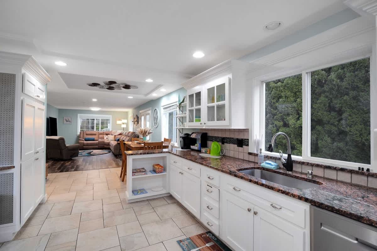 Modern kitchen with white shaker cabinets, granite countertops, and light tile flooring, featuring a large window overlooking greenery and an open view to the adjacent living area.