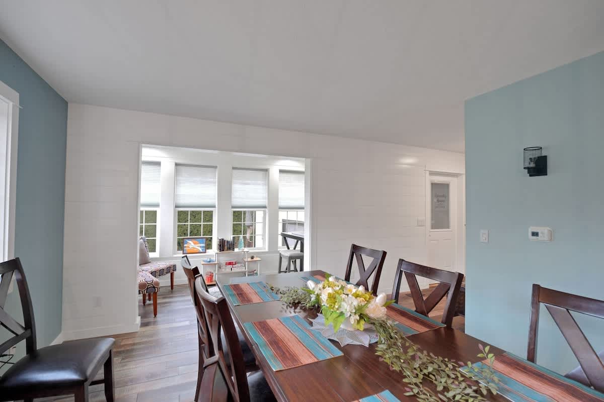 A bright dining area features a long wooden table with striped placemats and a floral centerpiece, set against a backdrop of white shiplap walls and a bay window with blinds.
