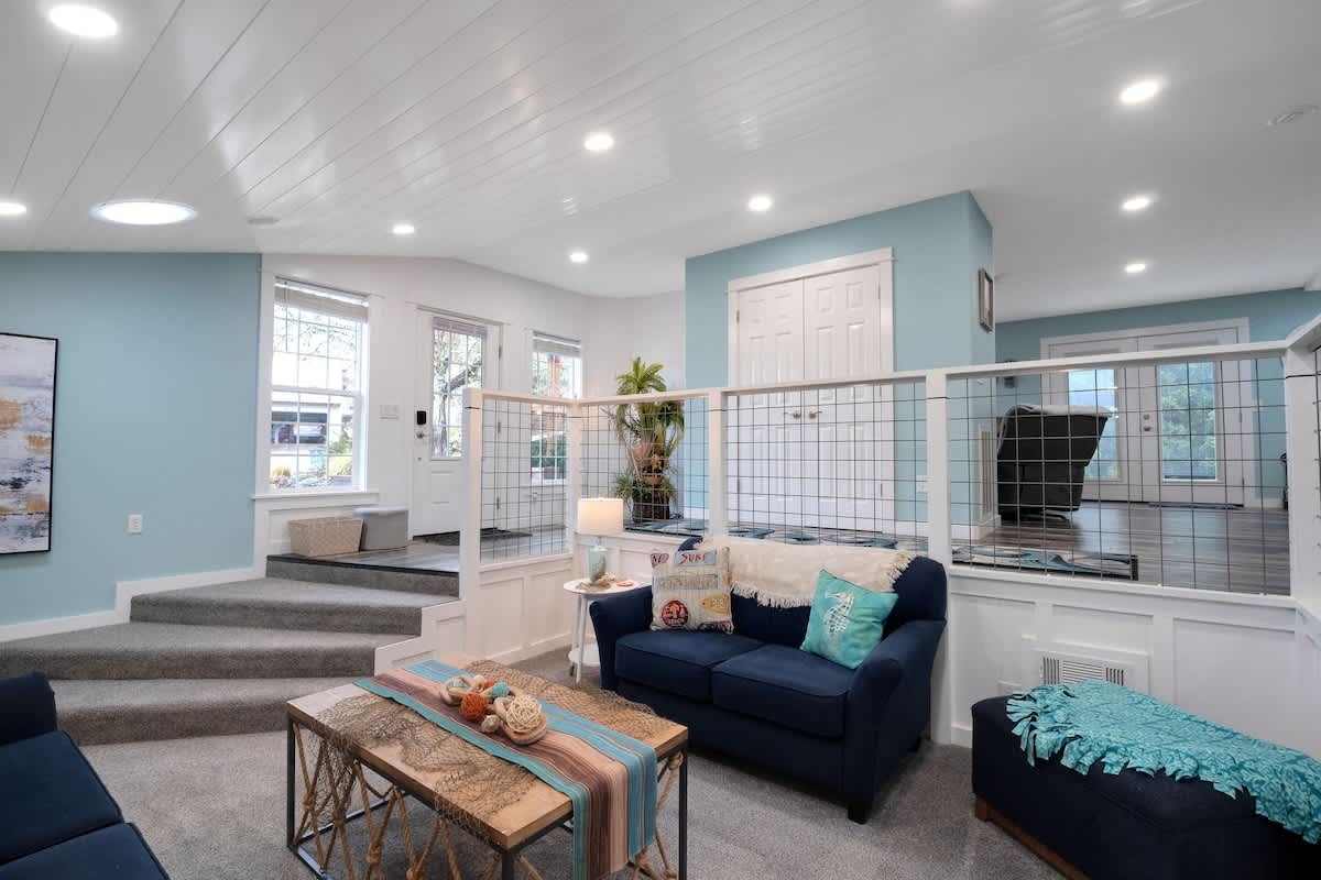 Expansive split-level living room with two navy blue sofas on plush gray carpet, facing a rustic coffee table, while an elevated landing with wood-look flooring and an entrance is visible above a white and wire mesh railing.