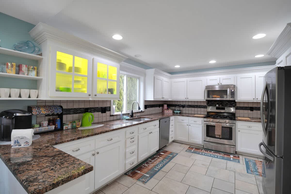 Bright kitchen with white cabinetry, dark granite countertops, and light tile flooring, featuring a stainless steel refrigerator, stove, and microwave, with a window above the sink.