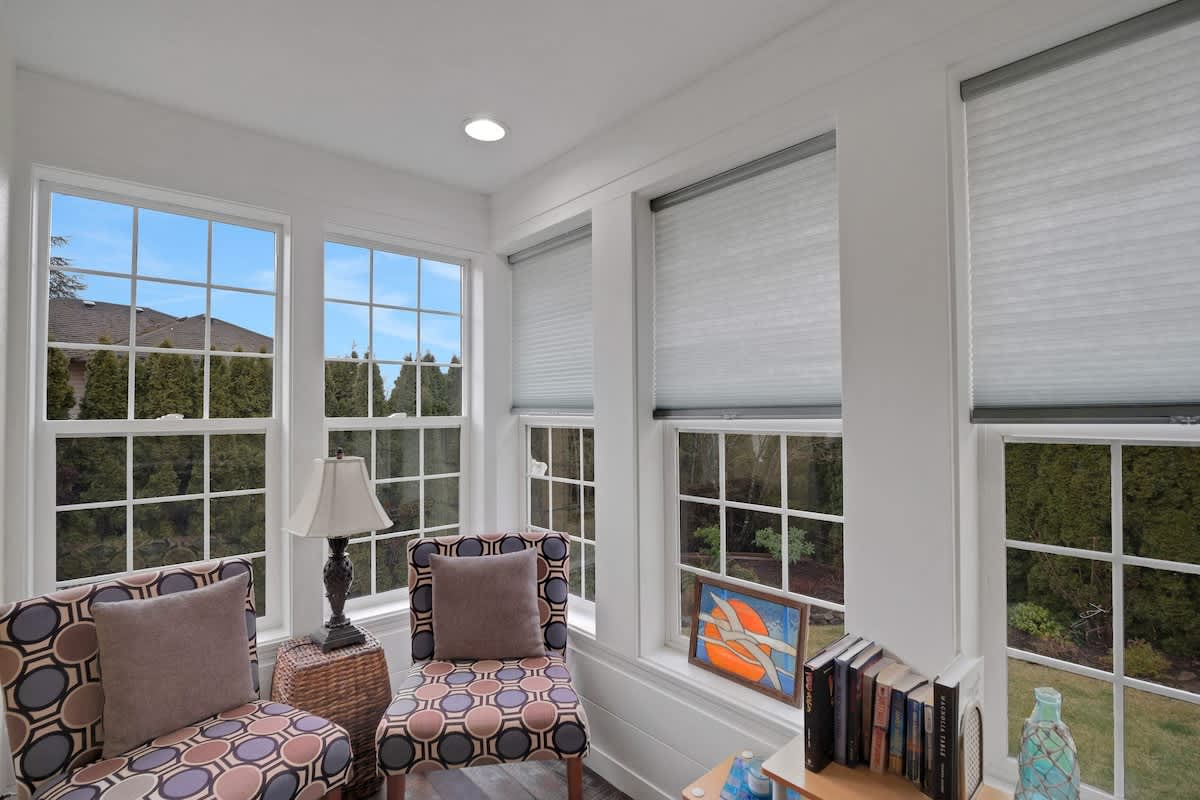 A bright sunroom or sitting area with white walls and numerous large windows offering views of the outdoors, furnished with two patterned armchairs and a small side table.