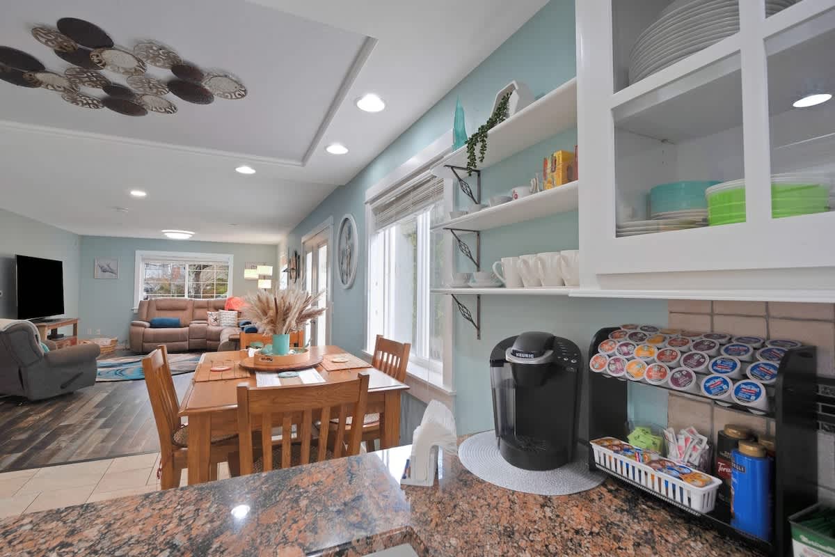 View from the kitchen into the dining area, featuring a wooden dining table with chairs, open shelving with dishes, and a coffee station, leading to a spacious living room beyond.