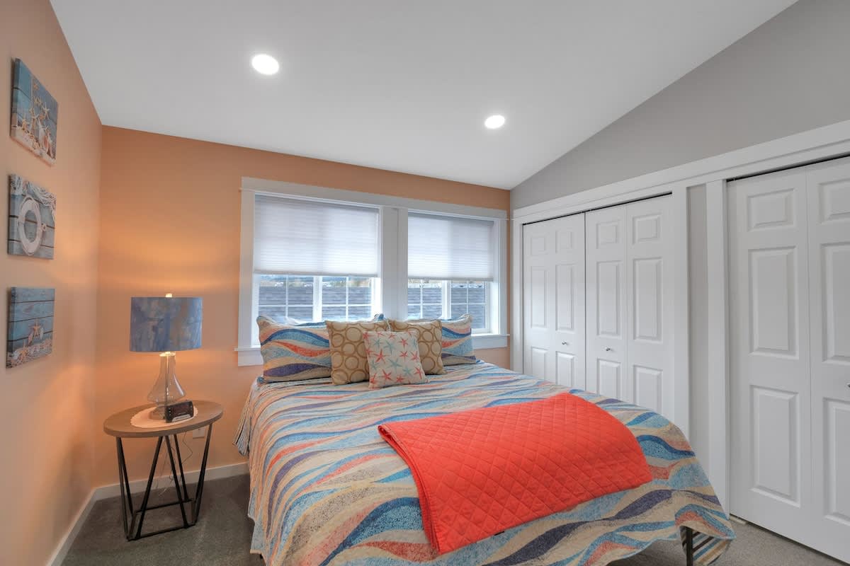This bedroom features a bed with a colorful striped quilt and an orange throw, a round nightstand with a blue lamp, and peach walls adorned with nautical-themed art. Windows with white blinds are visible.