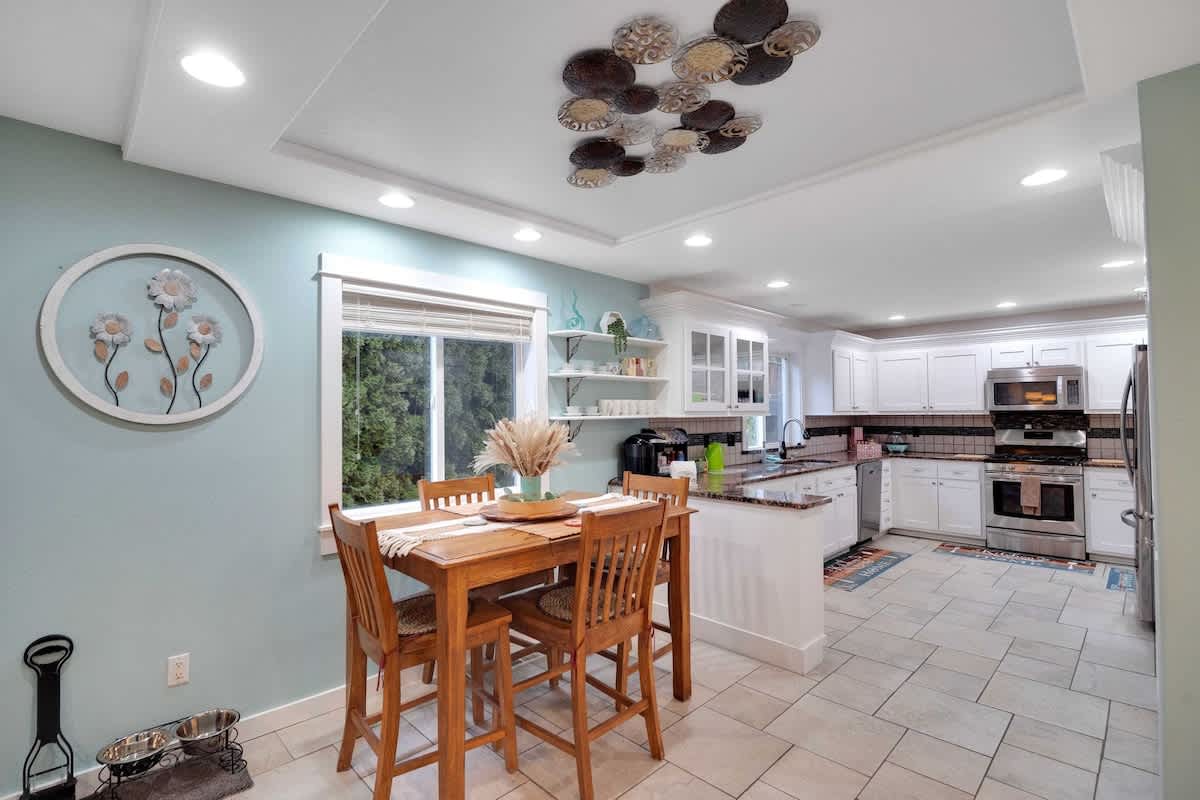 A bright kitchen with white shaker cabinets, stainless steel appliances, and light tile flooring, flowing into a dining area with a wooden high-top table and chairs. Light blue walls and decorative shelving add character.