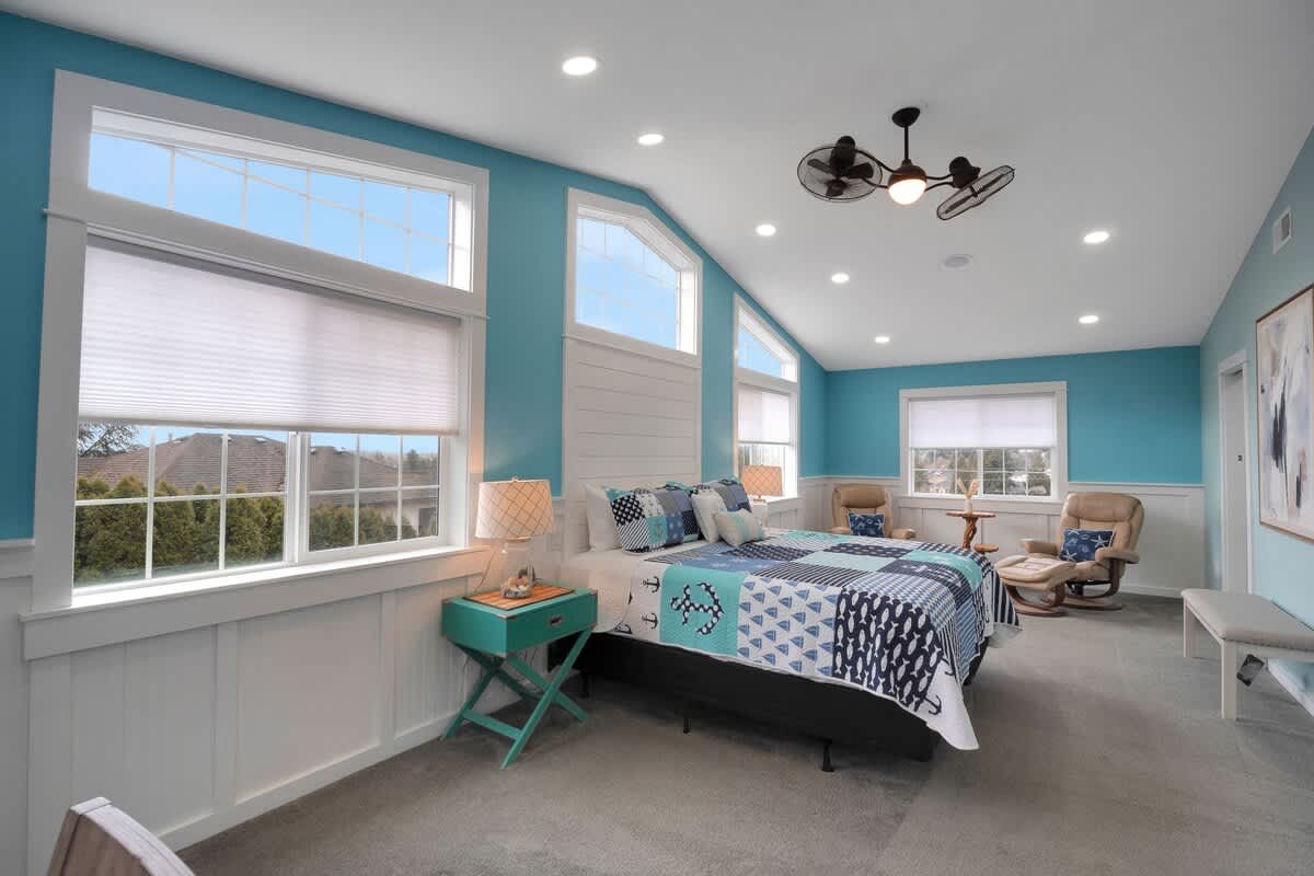 This wide shot of the light blue bedroom showcases the numerous large windows with white trim and blinds, a bed with a blue and white quilt, and a turquoise side table with a patterned lamp. Two armchairs are visible in the corner.
