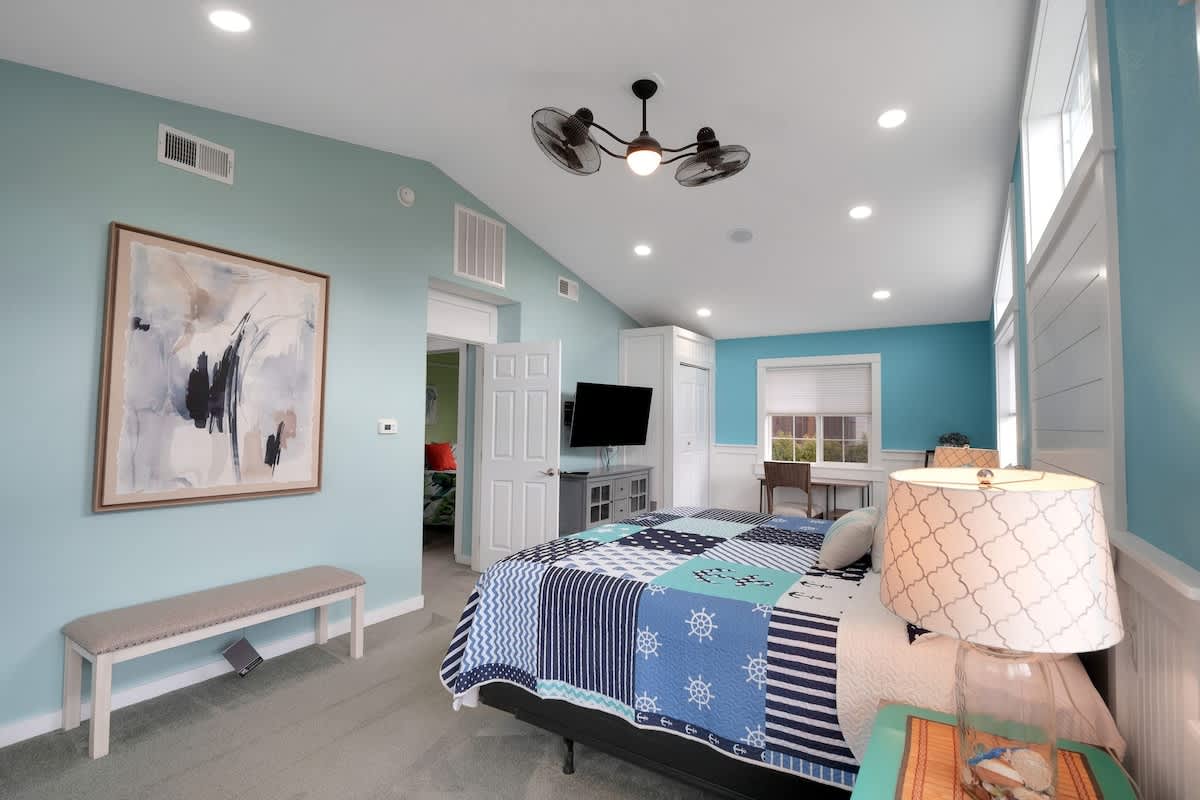 A spacious bedroom with light blue walls and white wainscoting, featuring a bed with a blue and white nautical patchwork quilt, a unique ceiling fan, and multiple large windows. Recessed lighting illuminates the room.