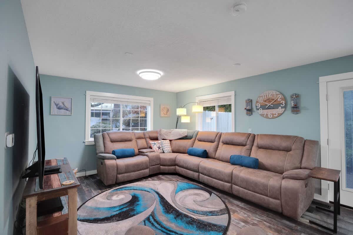 A cozy living room with light blue walls and dark wood-look flooring, featuring a large brown sectional sofa arranged around a round area rug, with a flat-screen TV on a rustic stand.