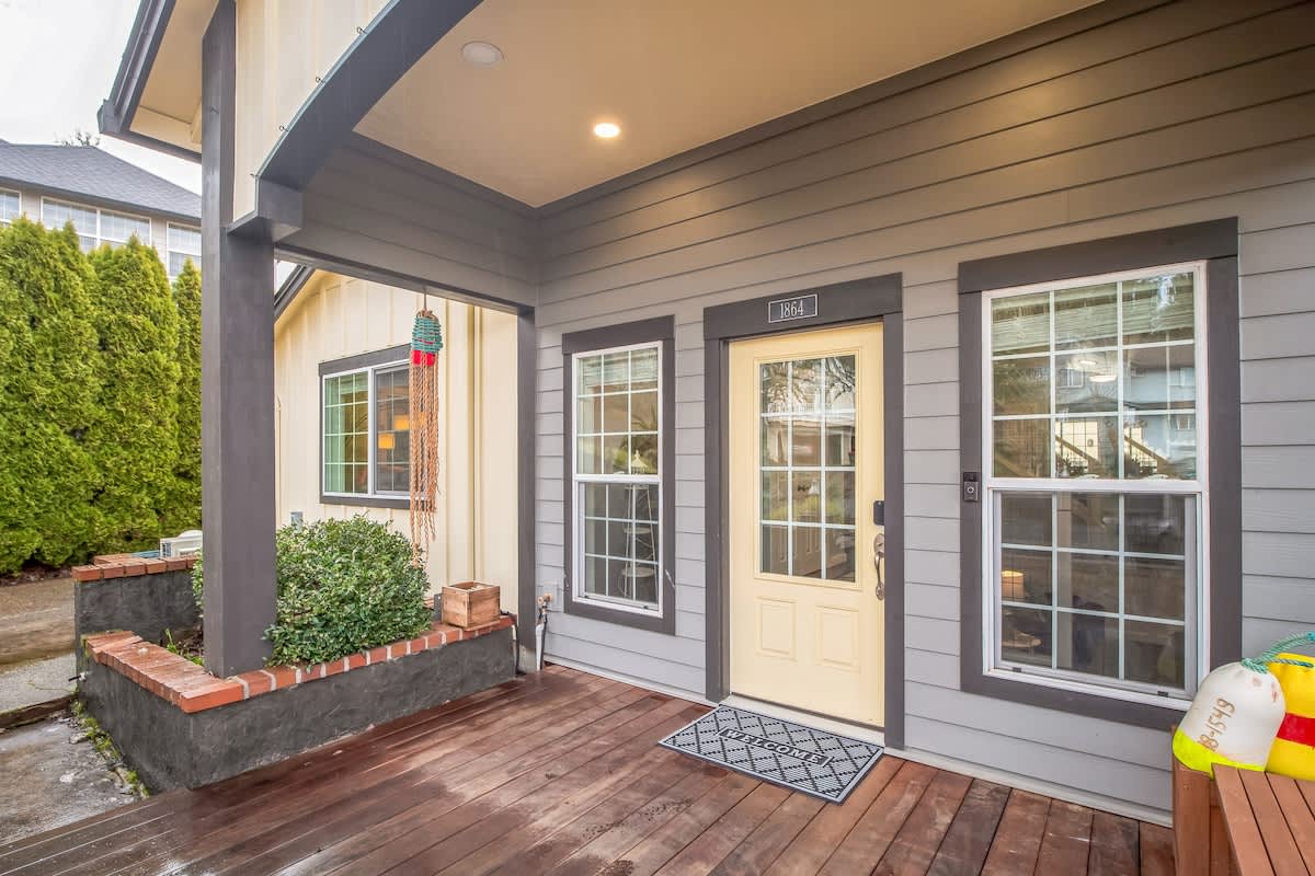 The front entrance of the house features a light yellow door with glass panes, flanked by windows and a small porch with a welcome mat. The siding is a mix of light yellow and gray.