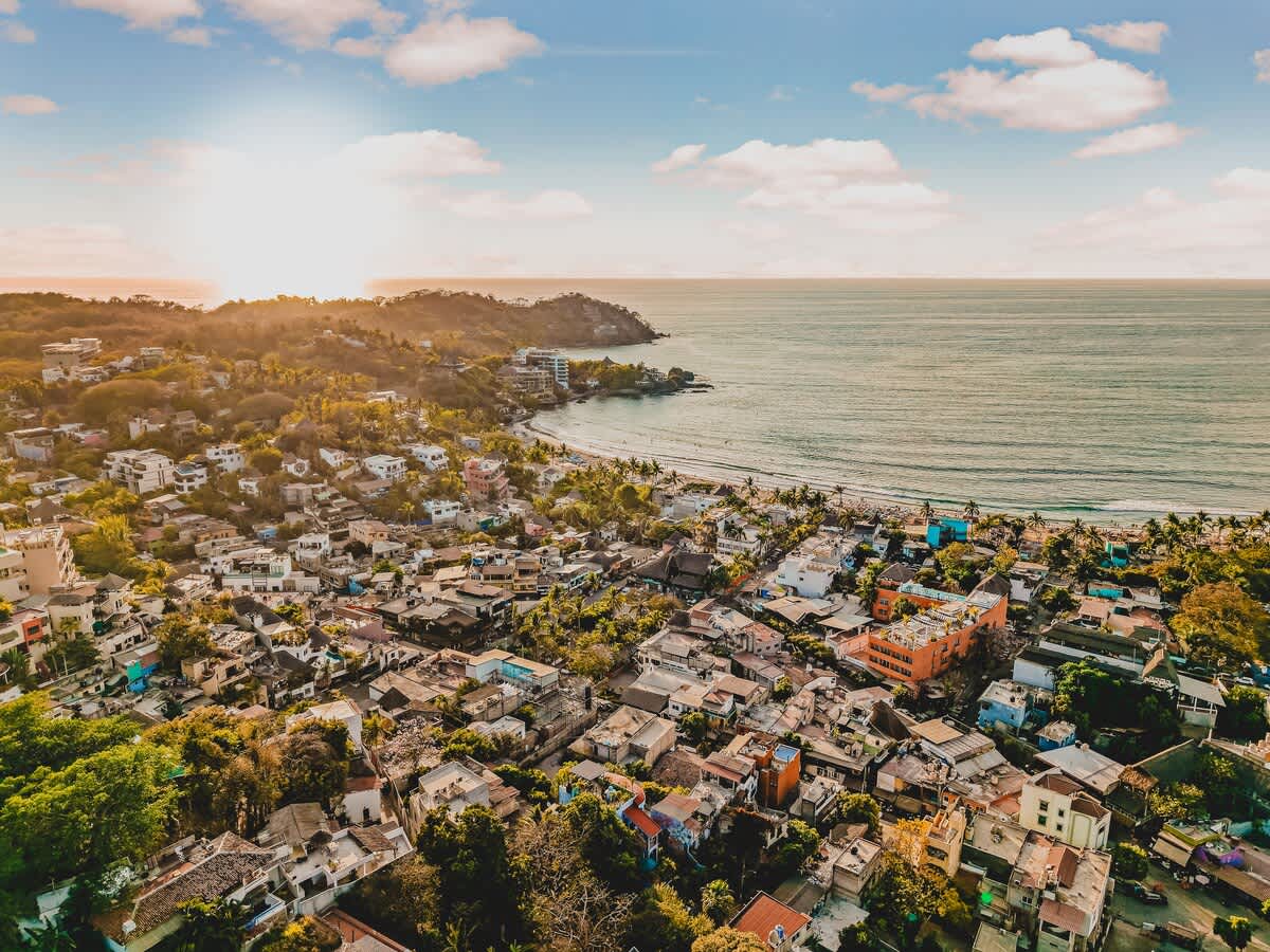 An aerial view captures a vibrant coastal town nestled between lush green hills and the sparkling ocean, with a sandy beach curving along the shoreline under a warm, sunlit sky.
