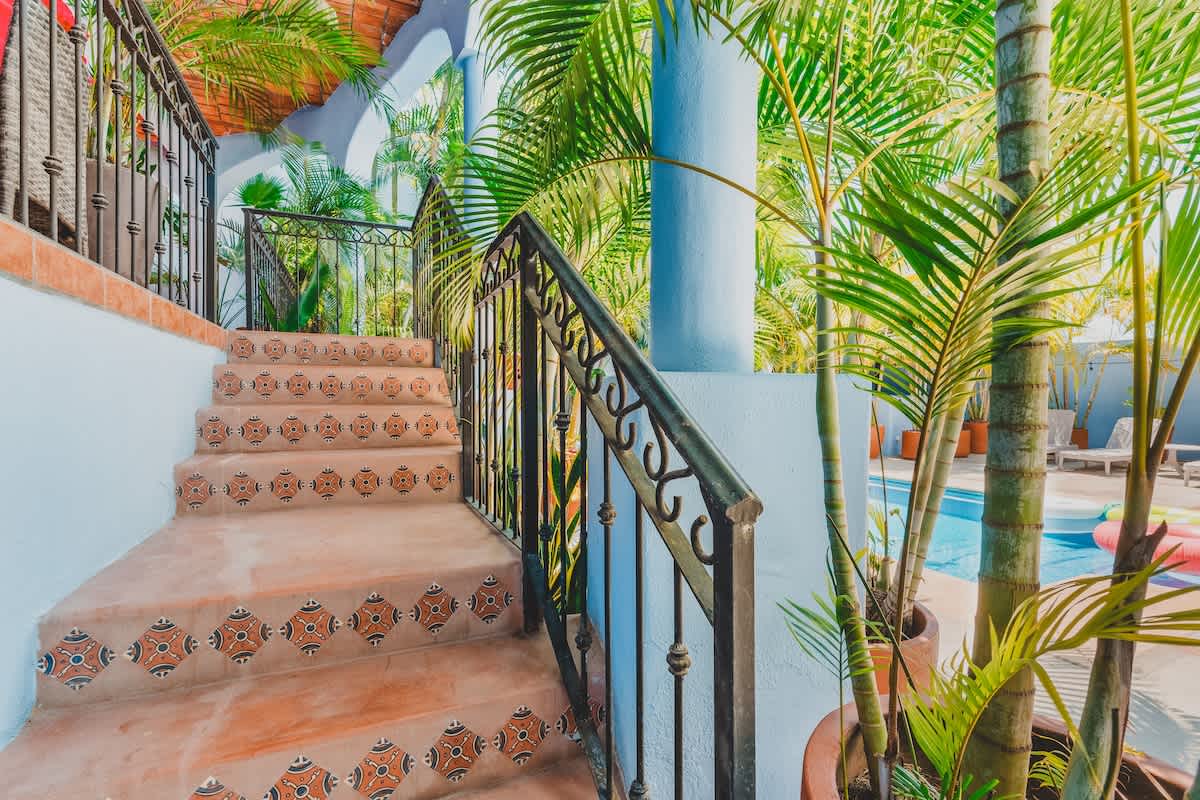 Outdoor staircase with decorative tiled risers and a black wrought-iron railing, leading down to a visible pool area surrounded by lush palm trees.