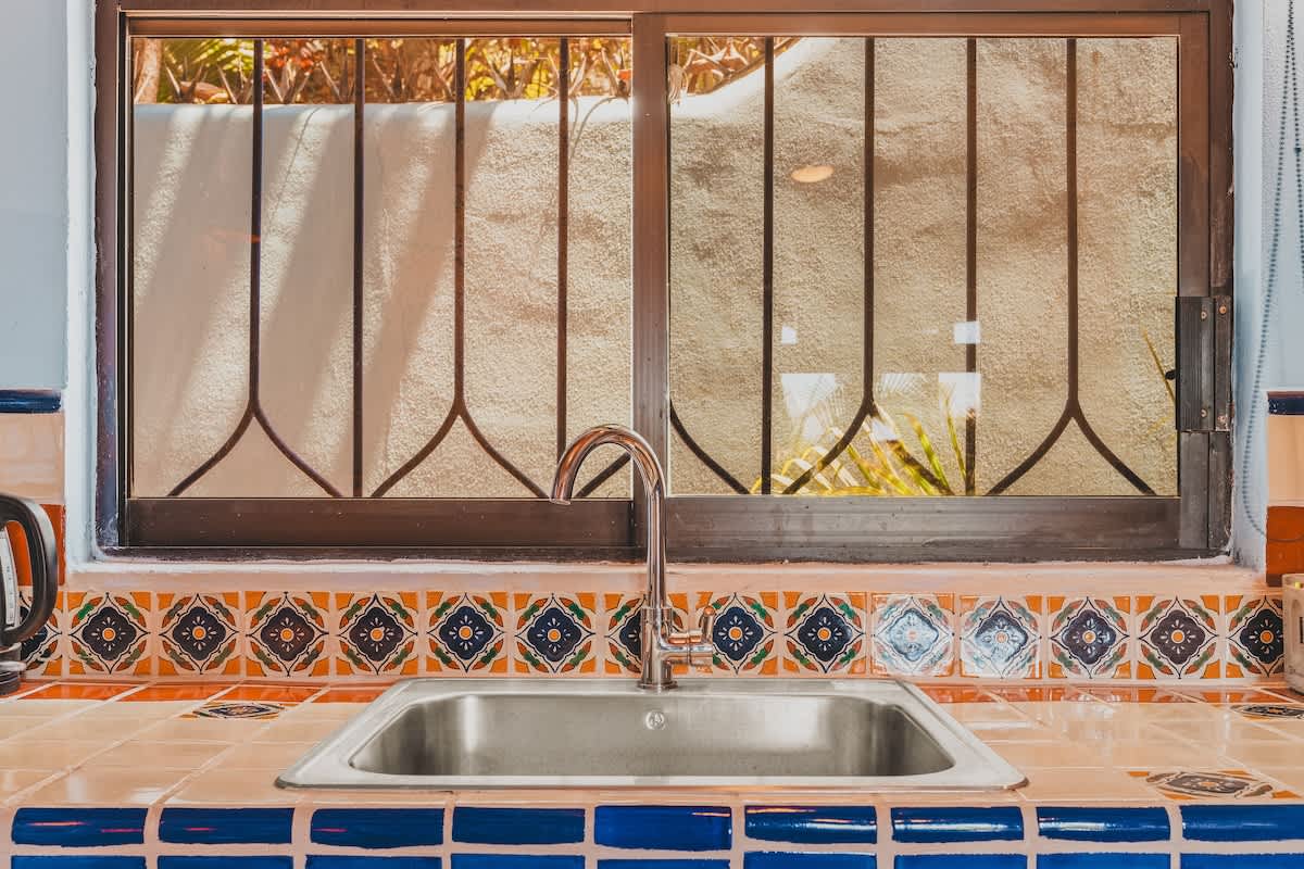 A close-up of the kitchen sink area with a stainless steel sink set into a colorful tiled countertop, featuring blue and orange decorative tiles, and a window with a metal security grill overlooking an outdoor area.