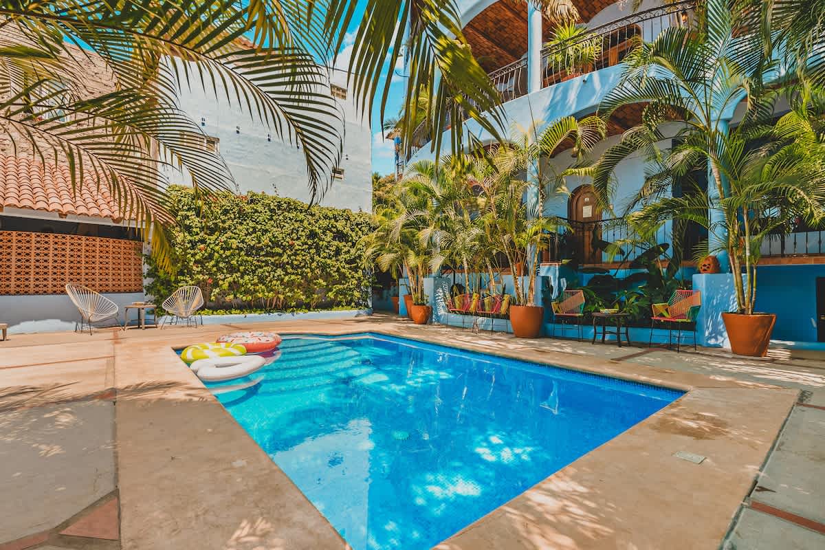 Inviting outdoor pool area with a rectangular blue-tiled pool, featuring colorful pool floats. The pool is bordered by concrete paving, and a light blue building with arched balconies and lush greenery provides a vibrant backdrop.