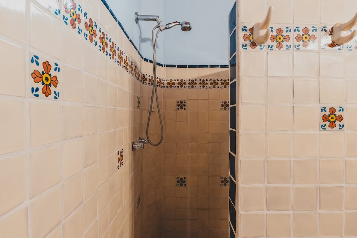 A shower stall with light beige square tiles and decorative floral accent tiles in orange and blue, featuring a wall-mounted shower head and a handheld shower, with a small step-up entrance.