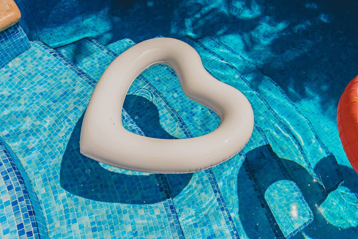 A close-up of the blue mosaic-tiled steps of a swimming pool, with a white heart-shaped inflatable float resting on the water.