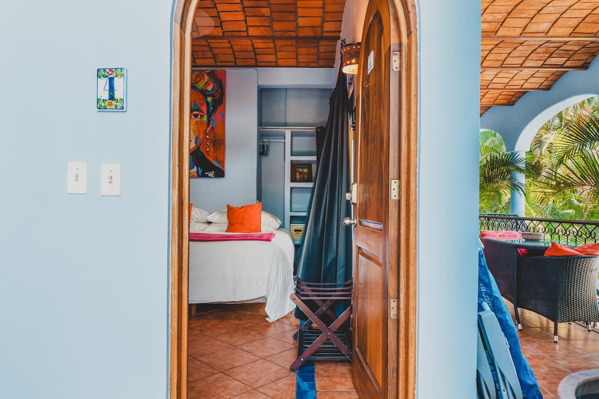 A view into the bedroom from an arched doorway, revealing the large bed with a pink blanket, a vibrant Frida Kahlo-inspired painting, and light blue walls with a terracotta tile ceiling. A luggage rack is positioned near the entrance, and a balcony with outdoor seating is visible through the open doors.