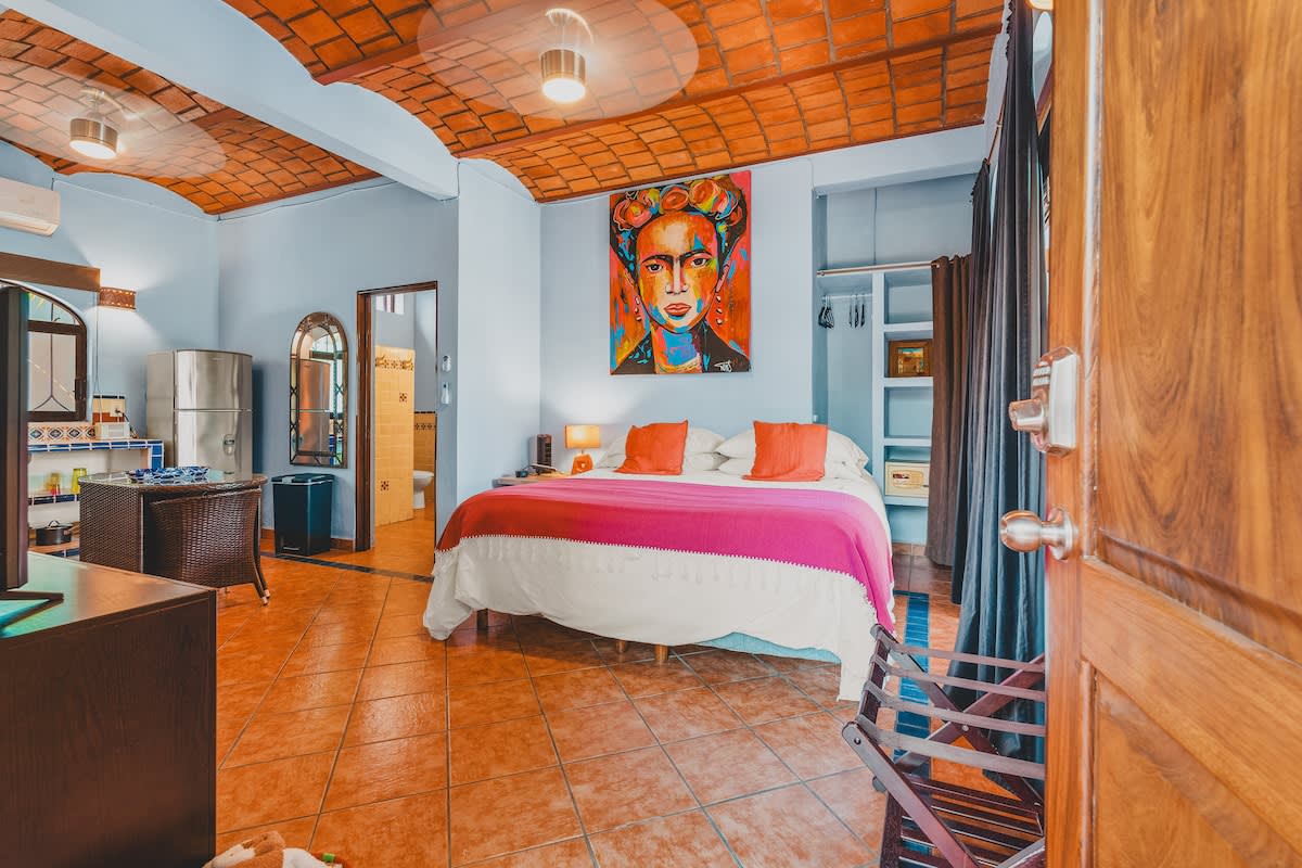 A wide shot of the colorful bedroom, showcasing the large bed with white linens and a pink and white blanket, a vibrant Frida Kahlo-inspired painting, and light blue walls with a terracotta tile ceiling. A small dining area with a glass-top table and wicker chairs is visible to the left, and a doorway leads to a bathroom.
