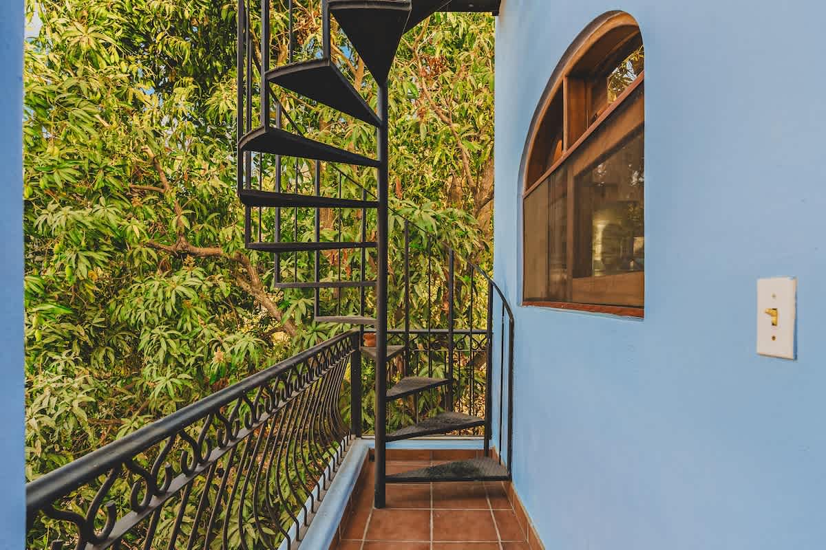 A black metal spiral staircase ascends from a tiled balcony, set against a light blue wall with an arched wooden window. Lush green trees provide a vibrant backdrop.