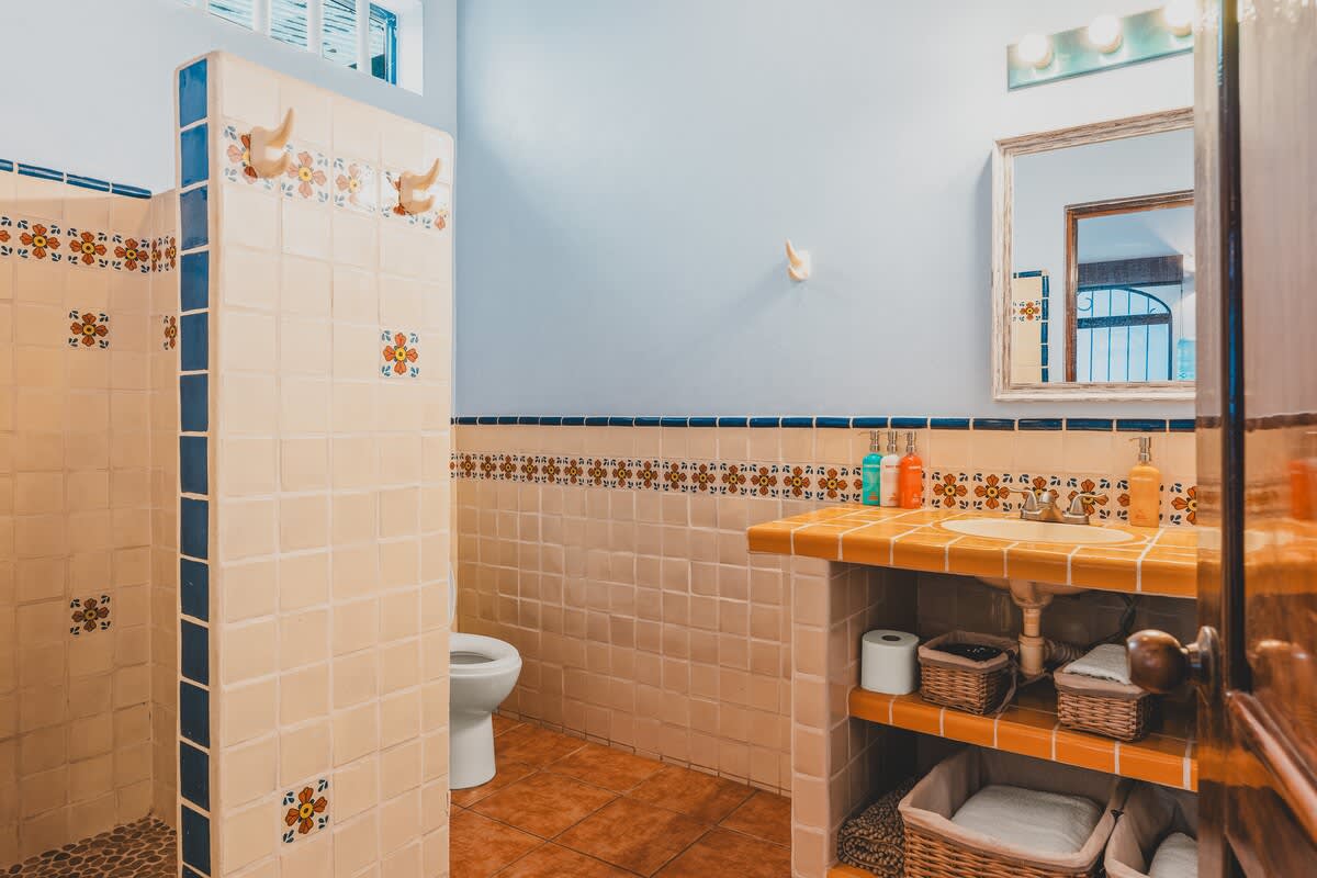 A bathroom with light beige tiled walls and a terracotta tiled floor, featuring a toilet, a tiled shower stall, and a vanity with a mirror, all accented with blue and orange floral decorative tiles.