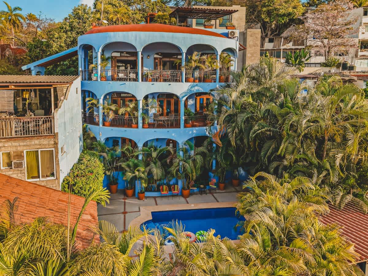Aerial view of the property, highlighting a multi-story light blue building with a red-tiled roof and numerous arched balconies adorned with potted plants. A bright blue swimming pool is visible in the courtyard, surrounded by lush tropical foliage and palm trees.