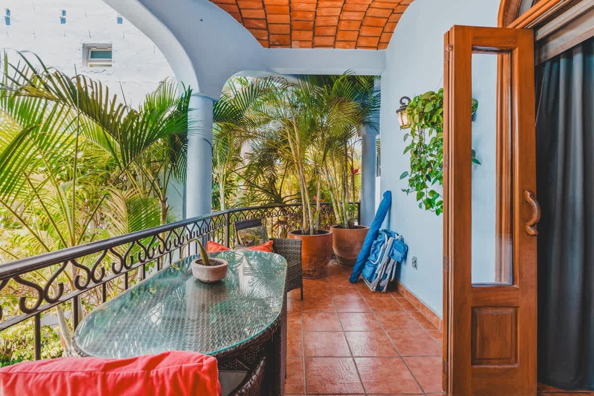 A wider view of the terracotta-tiled balcony with a curved, tiled ceiling, showing a wicker dining set and a sofa, surrounded by potted palm trees and lush greenery, with a blue backpack leaning against the wall.