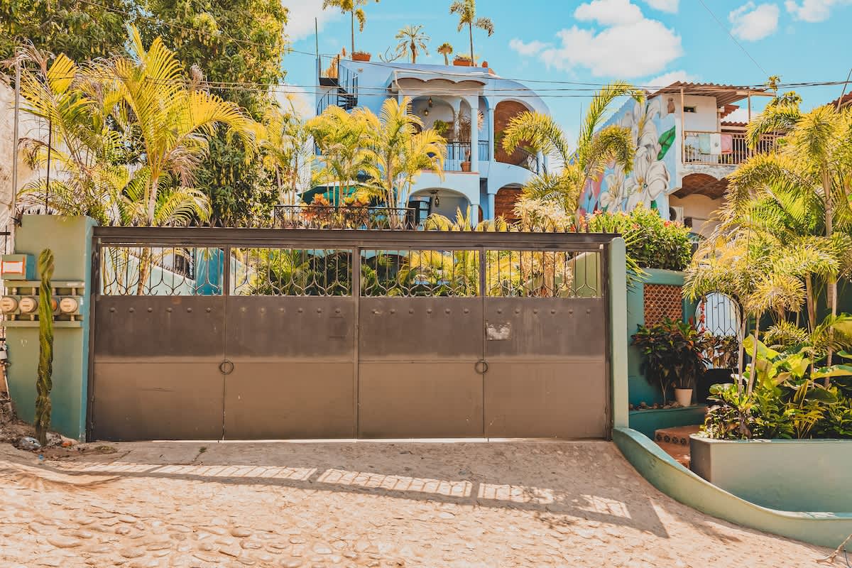 A large, dark metal gate with a decorative top section stands at the entrance of a property, fronting a cobblestone driveway. Behind it, a light blue multi-story building with balconies is visible amidst dense tropical greenery.