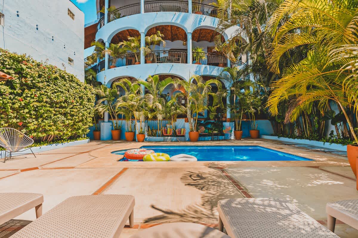 View of the pool area from behind lounge chairs, showing the bright blue pool with floats and the multi-story light blue building. The building features arched balconies and is framed by numerous potted palm trees and lush green foliage.