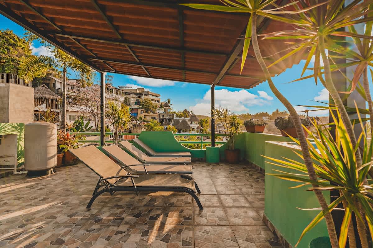 A spacious rooftop terrace featuring several lounge chairs on patterned tiled flooring, partially shaded by a large, open-sided canopy. Lush palm trees and tropical plants in pots add greenery, with a vibrant town and distant hills visible under a bright sky.