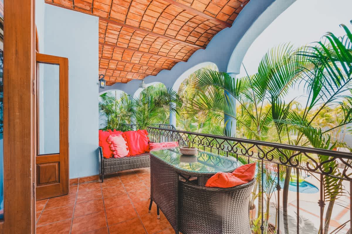 A vibrant balcony with terracotta tiled flooring and a curved, tiled ceiling, featuring a wicker sofa with bright red pillows and a glass-top dining table, overlooking lush palm trees and a distant pool.