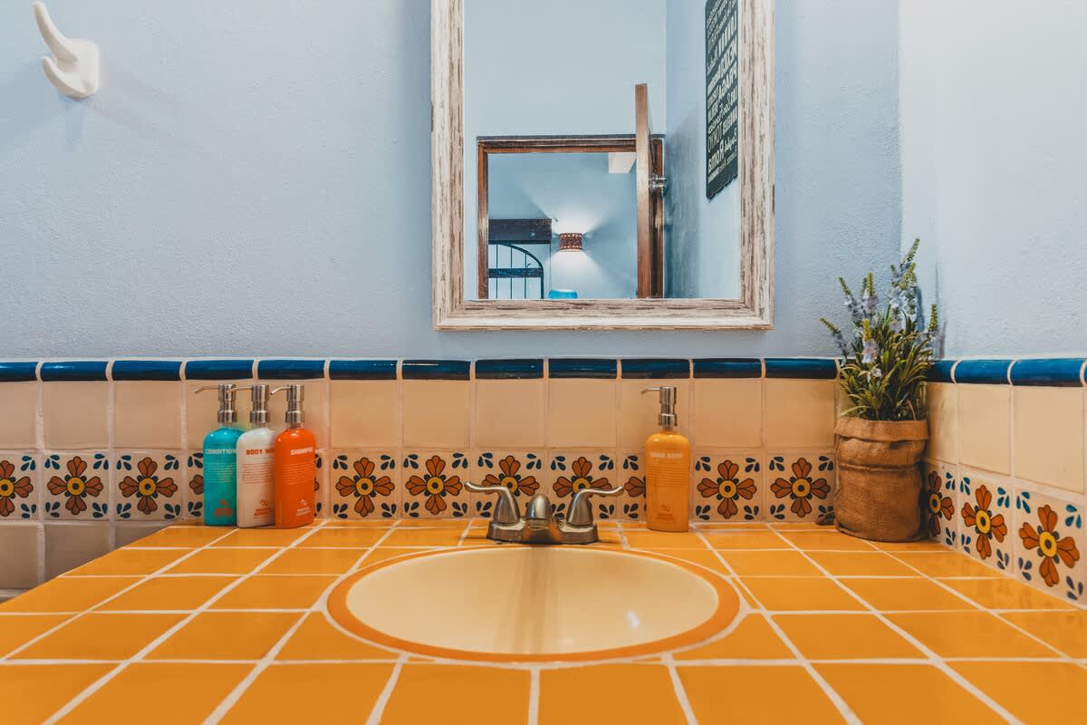 A close-up of the bathroom vanity with a yellow tiled countertop and an integrated sink, featuring a mirror above and a row of colorful toiletries, all set against a light blue wall with decorative floral tiles.