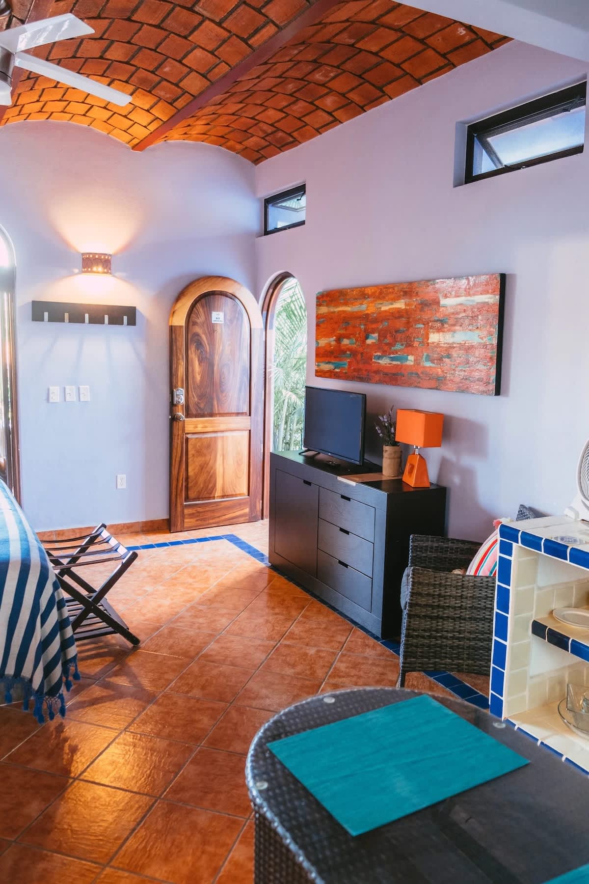 A view of the living area from a different angle, showing a dark wood dresser with a TV, an orange abstract painting, and a small dining table with a blue placemat. A wooden arched door leads outside.