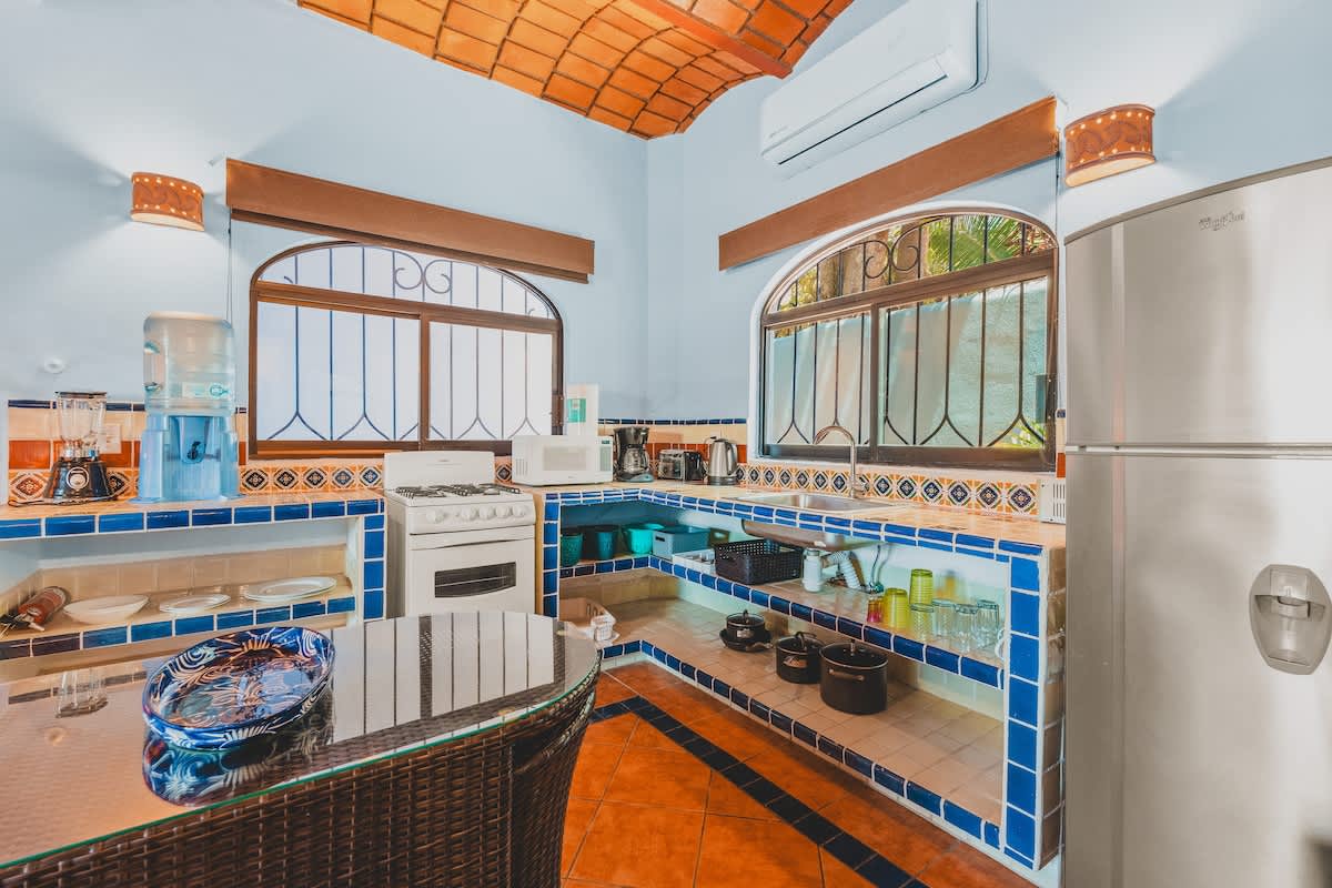 A vibrant kitchen featuring light blue walls, a vaulted terracotta tile ceiling, and blue and white tiled countertops and backsplash. It includes a white stove, microwave, refrigerator, and a glass-top dining table with wicker chairs.