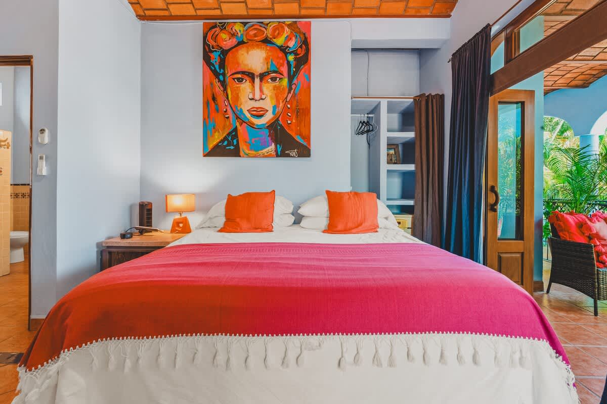 A close-up view of the bed in the colorful bedroom, featuring white linens, orange pillows, and a pink and white fringed blanket. A vibrant Frida Kahlo-inspired painting hangs above the bed, and a wooden nightstand with a lamp is beside it.