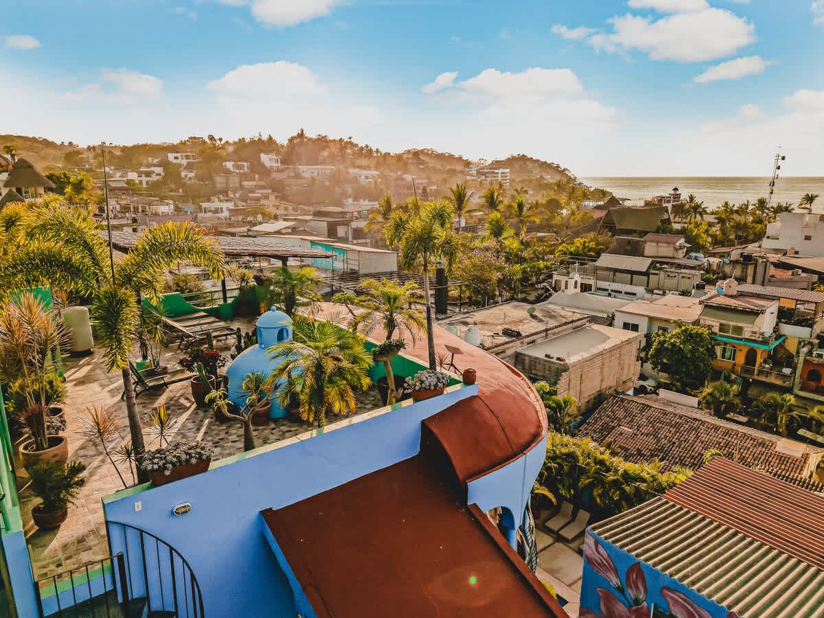 An expansive aerial view of the property's vibrant blue and terracotta rooftops, nestled within a lush tropical landscape overlooking a bustling coastal town and the ocean. Palm trees and distant hills complete the picturesque, sunny panorama.