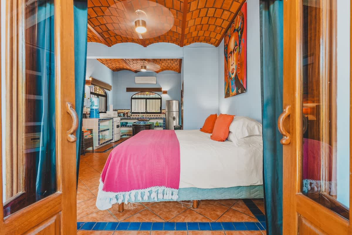 A view into the bedroom from the doorway, revealing a queen-sized bed with a white duvet and a bright pink throw, orange pillows, and a vibrant Frida Kahlo painting on the wall, under a vaulted brick ceiling.