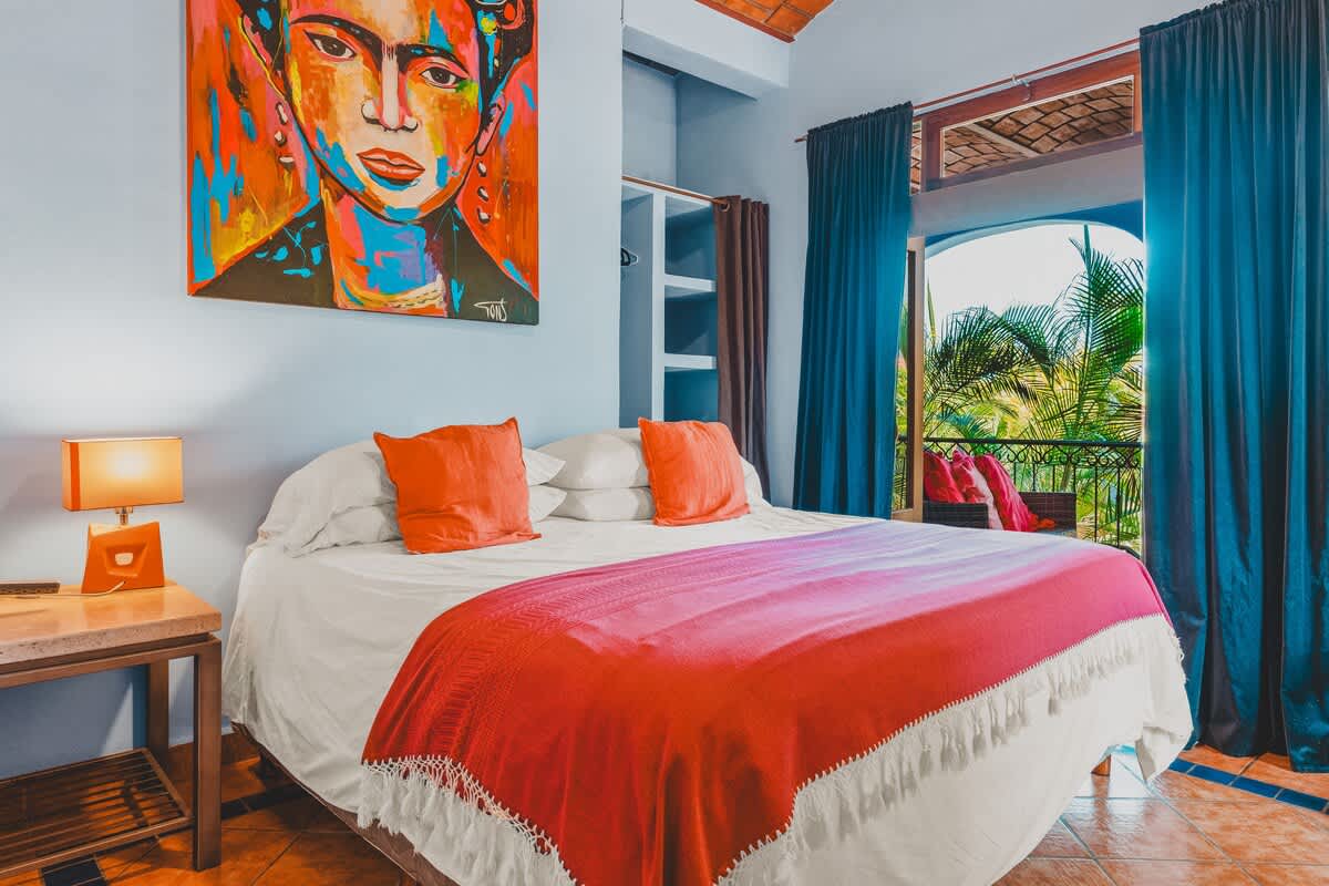A colorful bedroom with light blue walls, a terracotta tile ceiling, and a large bed with white linens, orange pillows, and a bright red blanket. A vibrant Frida Kahlo-inspired painting hangs above the bed, and blue curtains frame a doorway leading to a balcony with tropical foliage.