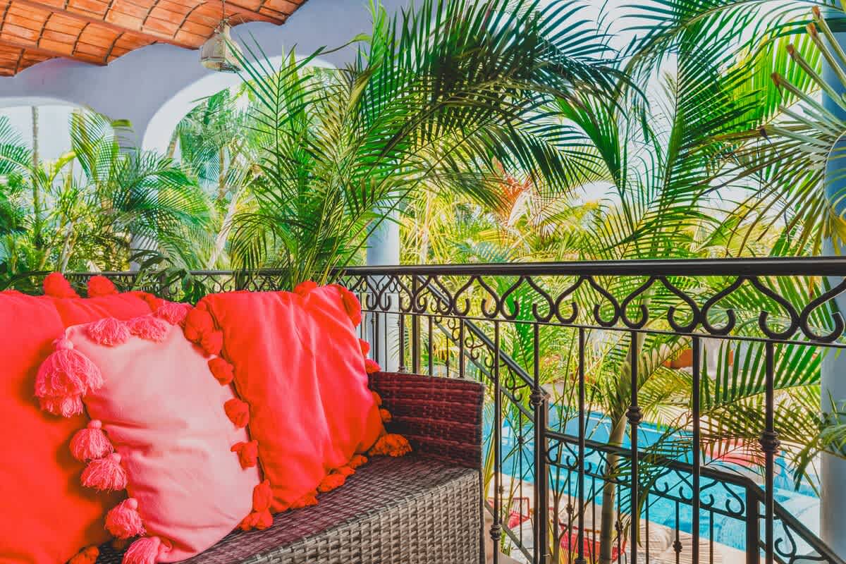 A close-up view of the balcony's wicker sofa adorned with bright red and pink tasseled pillows, set against a backdrop of vibrant green palm trees and a wrought iron railing, with a glimpse of a pool below.