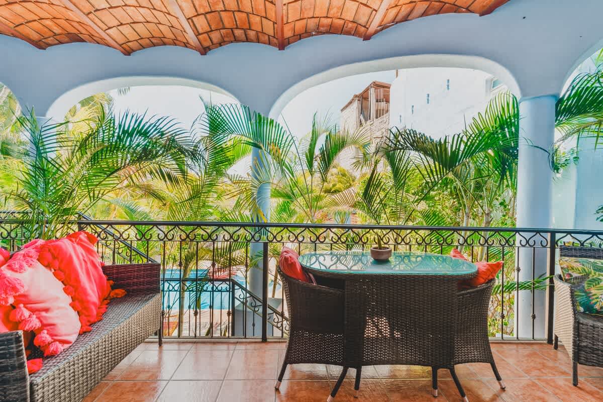 Covered balcony or patio with a glass-top dining table and wicker chairs, vibrant red cushions, and a wrought-iron railing, offering views of lush palm trees and a pool below.