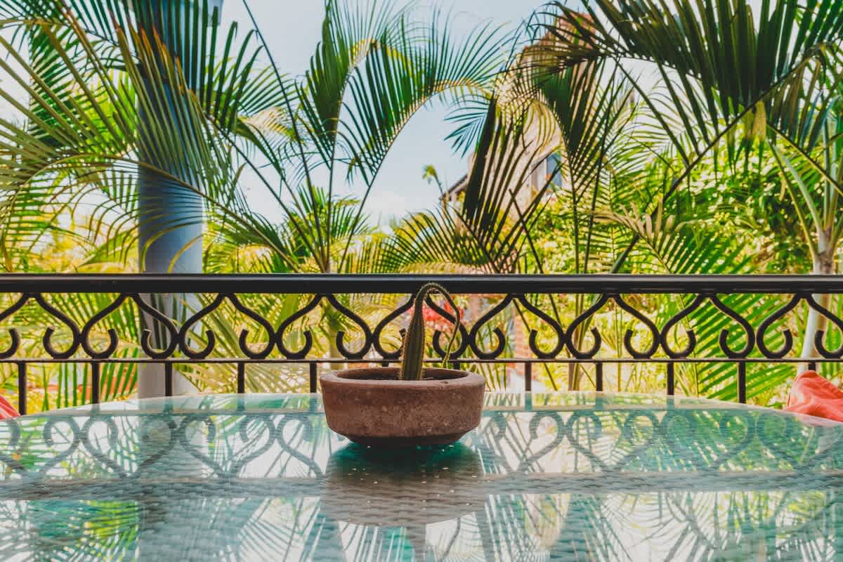 A detailed shot of the glass-top dining table on the balcony, reflecting the surrounding palm trees, with a small potted cactus as a centerpiece, and a decorative wrought iron railing in the foreground.