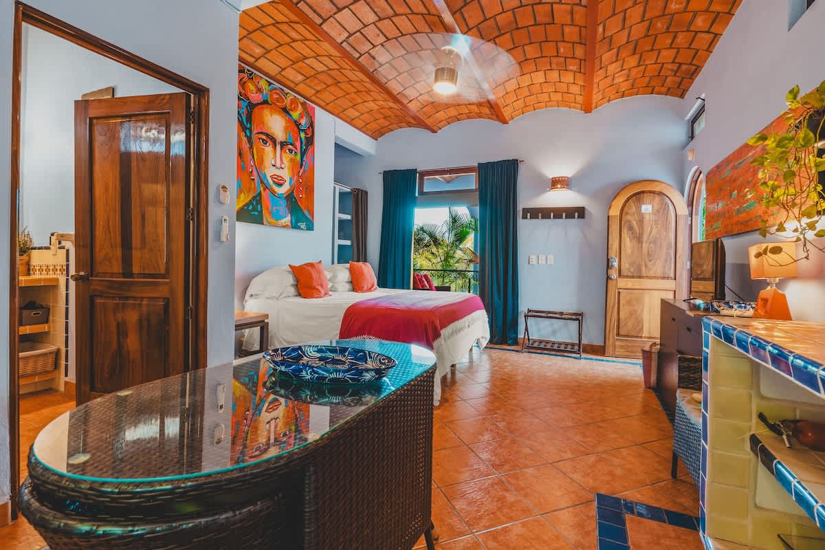 A spacious bedroom with terracotta tiled floors and a vaulted brick ceiling, featuring a queen bed with a red and white blanket, a large Frida Kahlo artwork, and a wicker dining table with a glass top.