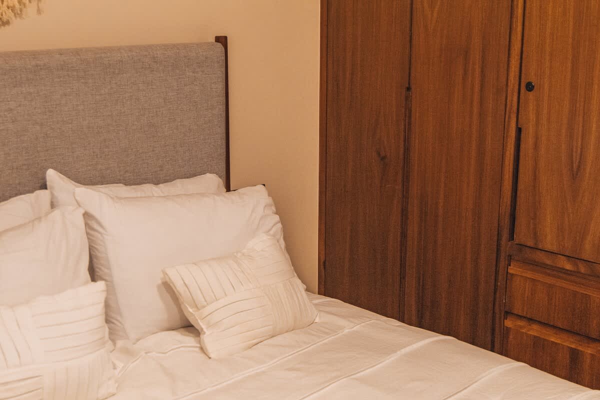A cozy bedroom corner featuring a bed with white bedding and a grey headboard, positioned next to a warm-toned wooden wardrobe with vertical paneling, suggesting ample storage.