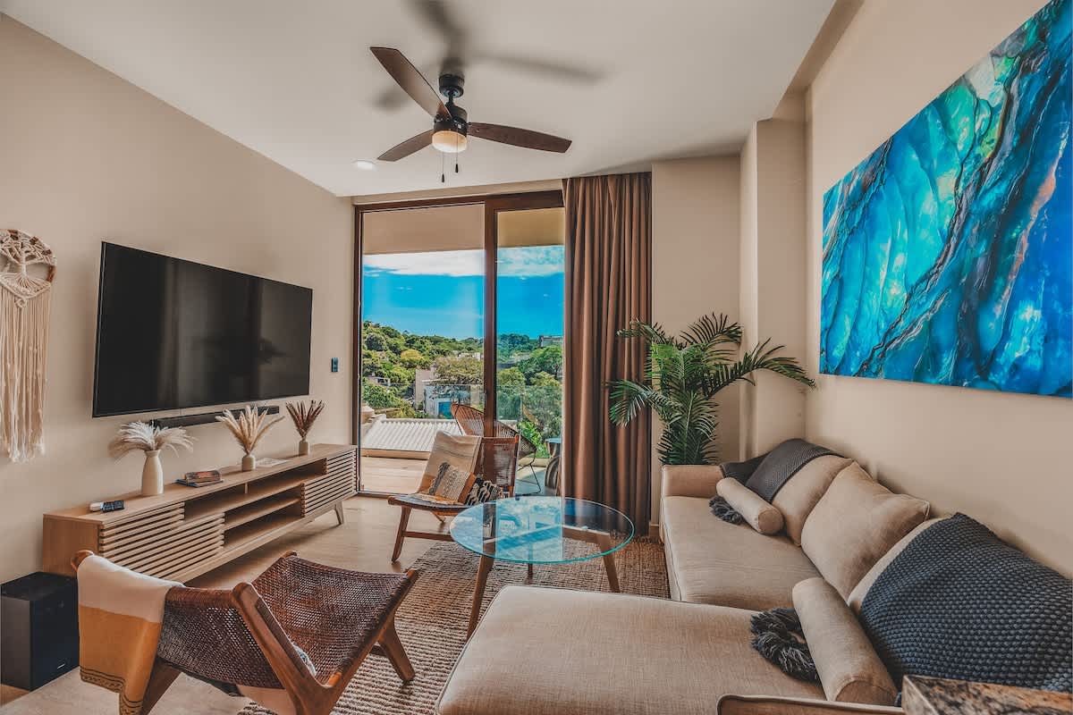 Bright living room with a light-colored L-shaped sectional sofa, a glass coffee table, and two woven chairs, centered around a wall-mounted TV and offering expansive views through a large sliding glass door.
