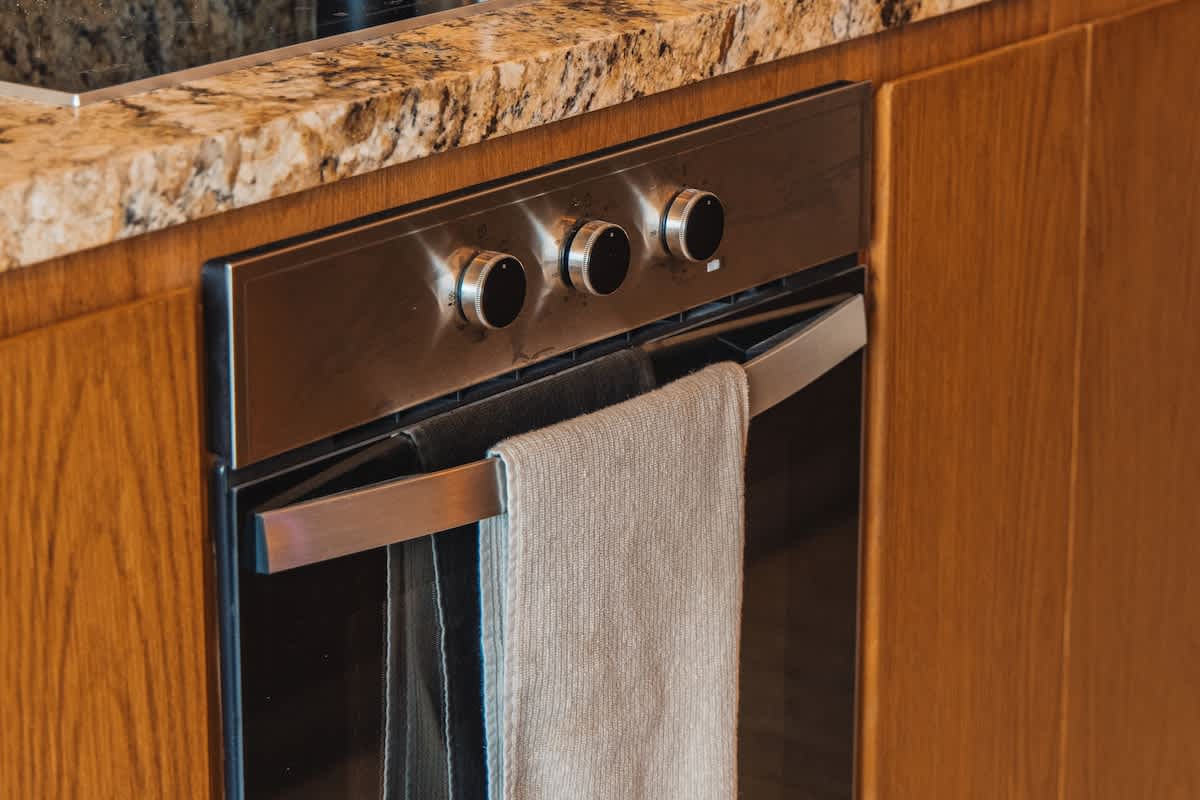 A modern stainless steel oven is integrated into rich wooden cabinetry, topped with a speckled granite countertop, with a light gray towel hanging from the oven handle.