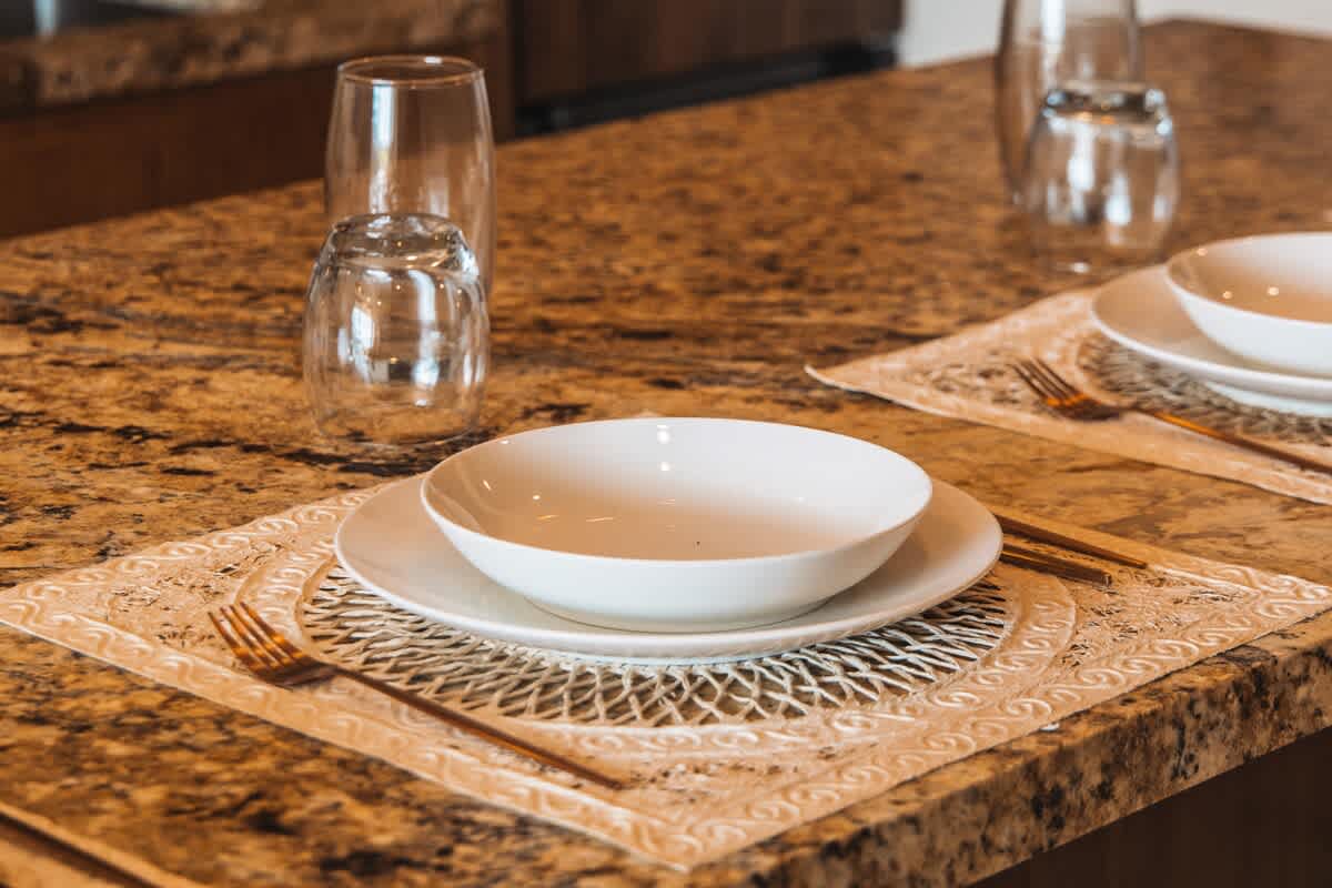 A place setting with white plates, a bowl, and gold-toned cutlery rests on a decorative placemat on a speckled granite countertop, with clear drinking glasses nearby.