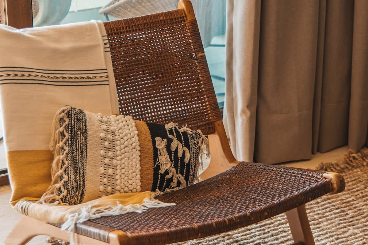 A stylish wooden lounge chair with woven cane details is adorned with a cream and yellow striped throw blanket and a textured decorative pillow featuring black, white, and gold patterns.
