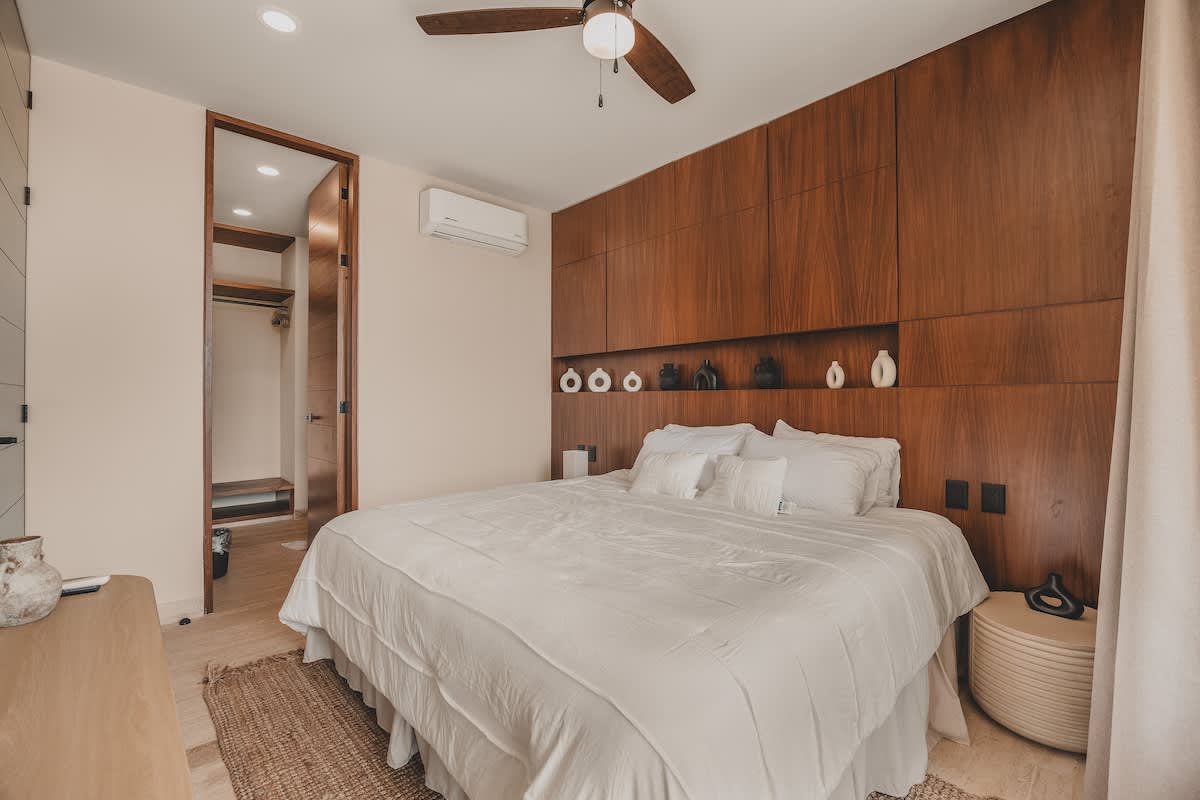 A view of the bedroom with a prominent wooden accent wall and integrated shelving behind a white-linened bed, featuring a round nightstand and a ceiling fan. An open doorway reveals a walk-in closet area.